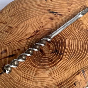 Puede incluir: Una broca de barrena de metal plateado con un diseño en espiral. La broca tiene una punta afilada y está sobre una superficie de madera con vetas visibles.