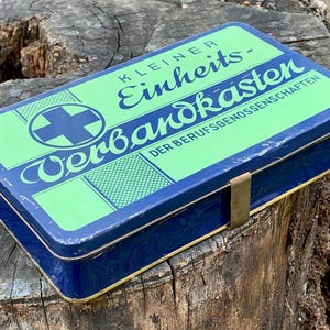 Op de afbeelding: Een vintage blauw en groen metalen EHBO-kistje. Het deksel is voorzien van de woorden "Kleiner Einheits-Verbandkasten" en een kruissymbool. De kist heeft een messing sluiting en staat op een verweerd houten oppervlak.