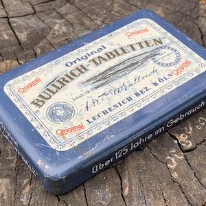 May include: A rectangular blue metal tin with the text "Original BULLRICH-TABLETTEN" in white lettering. The tin has a white and blue label with additional text and a cross symbol. The words "Über 125 Jahre im Gebrauch" are also visible.