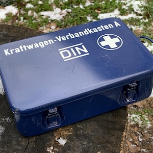 May include: A dark blue metal first aid kit with white text reading "Kraftwagen-Verbandkasten A" and "DIN". The kit features a white cross symbol and a handle. It is secured with two metal clasps.