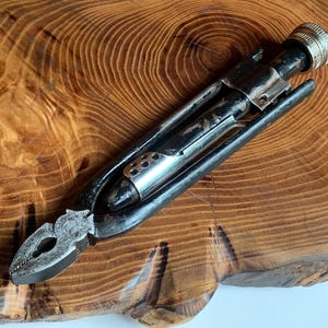 Peut inclure: Un outil multifonction en métal vintage doté d'une pince à une extrémité et d'un manche cylindrique avec un capuchon argenté moleté à l'autre. L'outil présente une finition noire avec des zones usées, une section centrale argentée et texturée.
