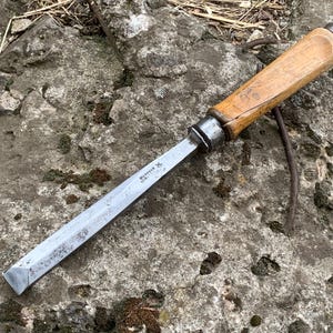 Puede incluir: Un formón de madera vintage con una hoja de metal. El formón tiene un mango de madera y una virola de metal. La hoja es plateada y tiene algo de texto. El formón está sobre una superficie de piedra rugosa y gris.