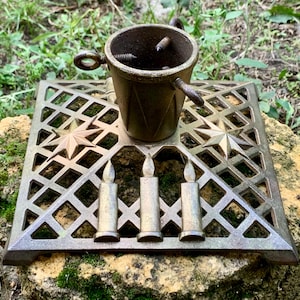 May include: A square, bronze-toned metal Christmas tree stand featuring a central cylindrical holder with three adjustable screws. The base has a decorative lattice pattern, two embossed eight-pointed stars, and three upright candle-shaped accents.