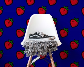 May include: A white chair with a grey blanket and black trainers on it. The chair is in front of a blue wall with a repeating pattern of red strawberries.