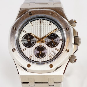 May include: A silver chronograph wristwatch with a white face and black subdials. The watch has a stainless steel band and a date window at the 4 o'clock position.