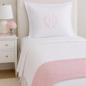 Puede incluir: Una funda de almohada blanca con una letra B bordada en rosa claro en el centro, rodeada por un dise&ntilde;o de corona de laurel. La cama est&aacute; cubierta con una colcha blanca acolchada y un camino de cama texturizado rosa claro.