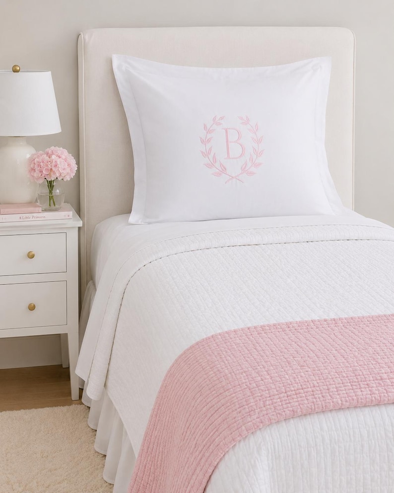 Puede incluir: Una funda de almohada blanca con una letra B bordada en rosa claro en el centro, rodeada por un dise&ntilde;o de corona de laurel. La cama est&aacute; cubierta con una colcha blanca acolchada y un camino de cama texturizado rosa claro.