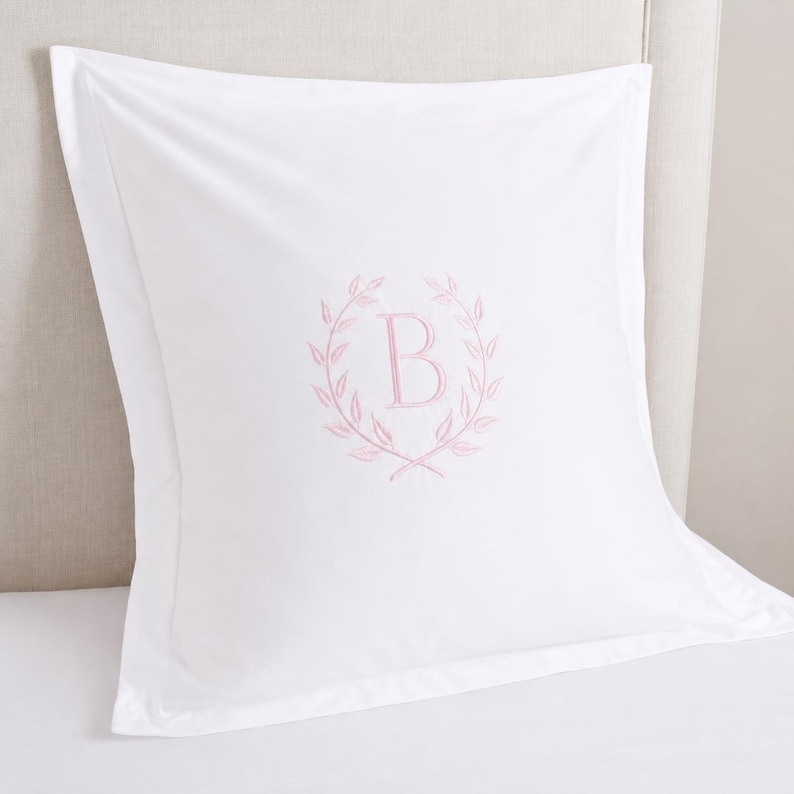 Puede incluir: Una funda de almohada cuadrada de color blanco con una letra B monogramada bordada en rosa claro en el centro, rodeada por un delicado dise&ntilde;o de corona de laurel. La funda tiene un borde ancho y un acabado de tela blanca lisa.