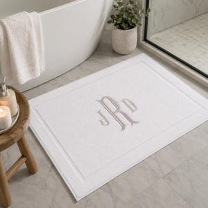 May include: A rectangular white bath mat featuring a centered, embroidered monogram in a light beige thread. The mat has a textured surface with a subtle border detail, providing a clean and simple aesthetic for a bathroom floor space.