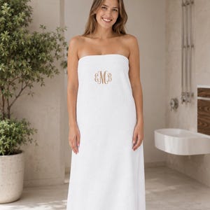 May include: A white strapless bath towel wrap featuring a centered, embroidered monogram in a light brown script font. The towel is made of a soft, absorbent terry cloth material and is designed to be worn around the torso for comfort and convenience.