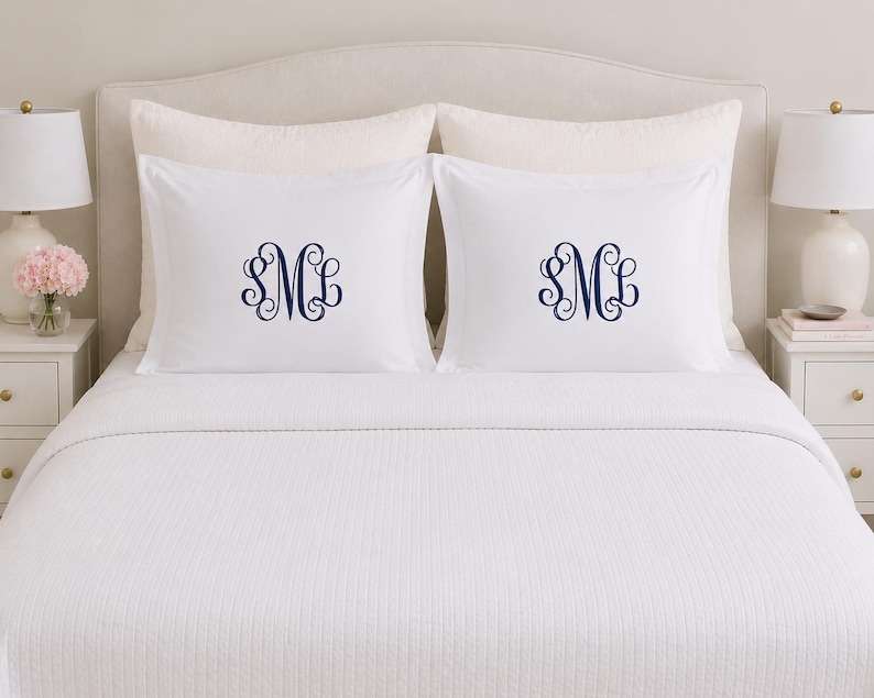 Puede incluir: Dos fundas de almohada rectangulares blancas con un monograma bordado en azul marino centrado con las letras SML en una fuente cursiva decorativa. Las fundas est&aacute;n colocadas sobre una colcha blanca con un patr&oacute;n texturizado.