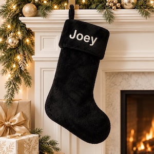 May include: A black plush Christmas stocking featuring a folded cuff at the top. The name Joey is embroidered in white capital and lowercase letters on the center of the cuff. The stocking has a soft, fuzzy texture and a hanging loop at the top.