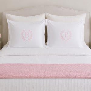 Puede incluir: Un juego de cama blanco con dos fundas de almohada que presentan una letra B bordada en rosa claro rodeada por una corona de laurel. La cama est&aacute; cubierta con un edred&oacute;n blanco acolchado y un camino de cama acolchado rosa claro liso.