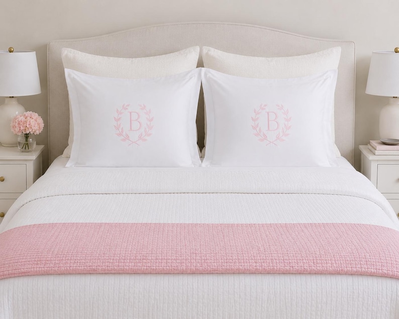 Puede incluir: Un juego de cama blanco con dos fundas de almohada que presentan una letra B bordada en rosa claro rodeada por una corona de laurel. La cama est&aacute; cubierta con un edred&oacute;n blanco acolchado y un camino de cama acolchado rosa claro liso.