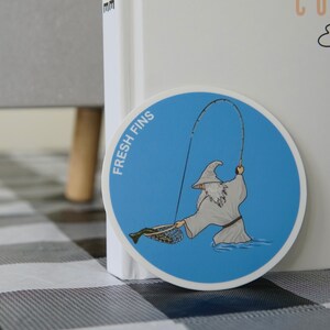 Puede incluir: Una pegatina azul y blanca con un dibujo animado de un mago pescando. La pegatina dice "Fresh Fins" y presenta a un mago con una t&uacute;nica blanca sosteniendo una ca&ntilde;a de pescar y una red con un pez dentro.