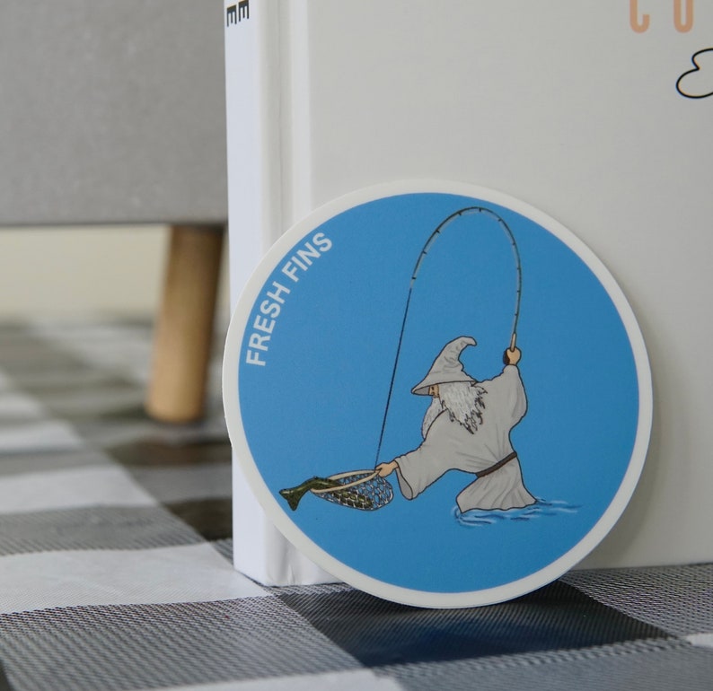 Puede incluir: Una pegatina azul y blanca con un dibujo animado de un mago pescando. La pegatina dice "Fresh Fins" y presenta a un mago con una t&uacute;nica blanca sosteniendo una ca&ntilde;a de pescar y una red con un pez dentro.