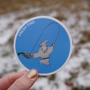 Puede incluir: Una pegatina redonda azul con un borde blanco. La pegatina presenta una ilustraci&oacute;n de dibujos animados de una persona con una t&uacute;nica de mago sosteniendo una ca&ntilde;a de pescar. El texto "FRESH FINS" est&aacute; escrito en blanco en la pegatina.