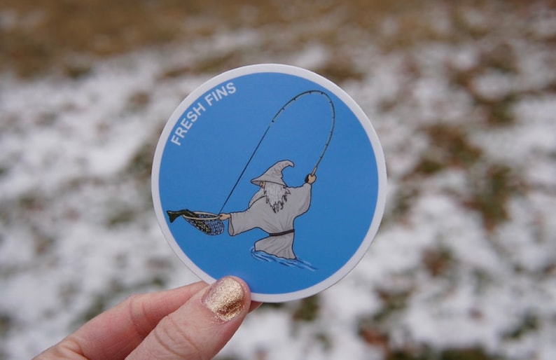 Puede incluir: Una pegatina redonda azul con un borde blanco. La pegatina presenta una ilustraci&oacute;n de dibujos animados de una persona con una t&uacute;nica de mago sosteniendo una ca&ntilde;a de pescar. El texto "FRESH FINS" est&aacute; escrito en blanco en la pegatina.