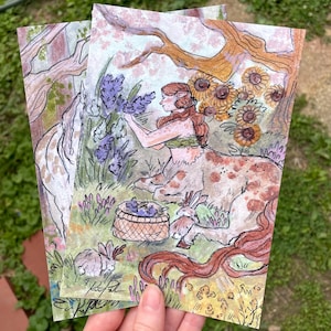Op de afbeelding: Twee kunstdrukken met een fantasie-illustratie van een centaurachtig wezen dat lavendel plukt. Het kunstwerk bevat een mand met bessen, konijnen en zonnebloemen, in een boslandschap. De prints worden in een hand gehouden tegen een groene achtergrond.