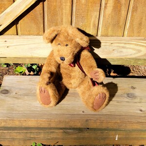 May include: A brown teddy bear with a dark red ribbon around its neck. The bear has a black nose and brown paws. It is sitting on a wooden bench. The bear is a classic toy.