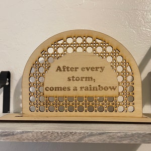 May include: A wooden sign with a woven pattern in the shape of an arch. The sign says "After every storm, comes a rainbow".