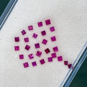 Könnte beinhalten: Eine Ansammlung kleiner, quadratisch geschliffener, leuchtend magentafarbener Edelsteine, die auf einer weißen Oberfläche in einem transparenten Behälter angeordnet sind. Die Edelsteine sind gleichmäßig groß und reflektieren das Licht.