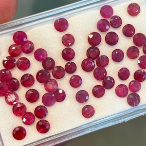 May include: A collection of ruby red gemstones in a clear plastic box. The gemstones are round and faceted.