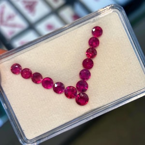 Pode incluir: Um close-up de uma pequena caixa de plástico transparente contendo uma coleção de 15 pedras preciosas redondas de cor rosa brilhante. As pedras preciosas estão dispostas em forma de V.