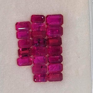 Puede incluir: Una colección de 15 piedras preciosas rectangulares de color rosa vibrante, probablemente rubíes, dispuestas en filas sobre una superficie blanca.