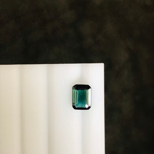 May include: A dark green gemstone with a rectangular cut, set on a white surface.