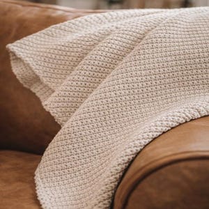 May include: A folded, beige crocheted blanket draped over a brown leather couch. The blanket has a textured, woven appearance and is folded to reveal multiple layers. The neutral color palette and soft texture suggest comfort and warmth.