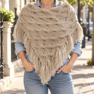 Peut inclure: Un poncho tricoté beige avec un motif en losanges texturé et de longues franges sur les bords inférieurs. Le vêtement est porté sur une chemise boutonnée bleu clair et un jean bleu, créant un look superposé avec un ourlet en pointe.