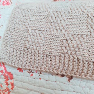 May include: Close-up of a beige knitted blanket with a textured pattern. The blanket is made of thick yarn and has a geometric design. The blanket is resting on a floral patterned quilt.