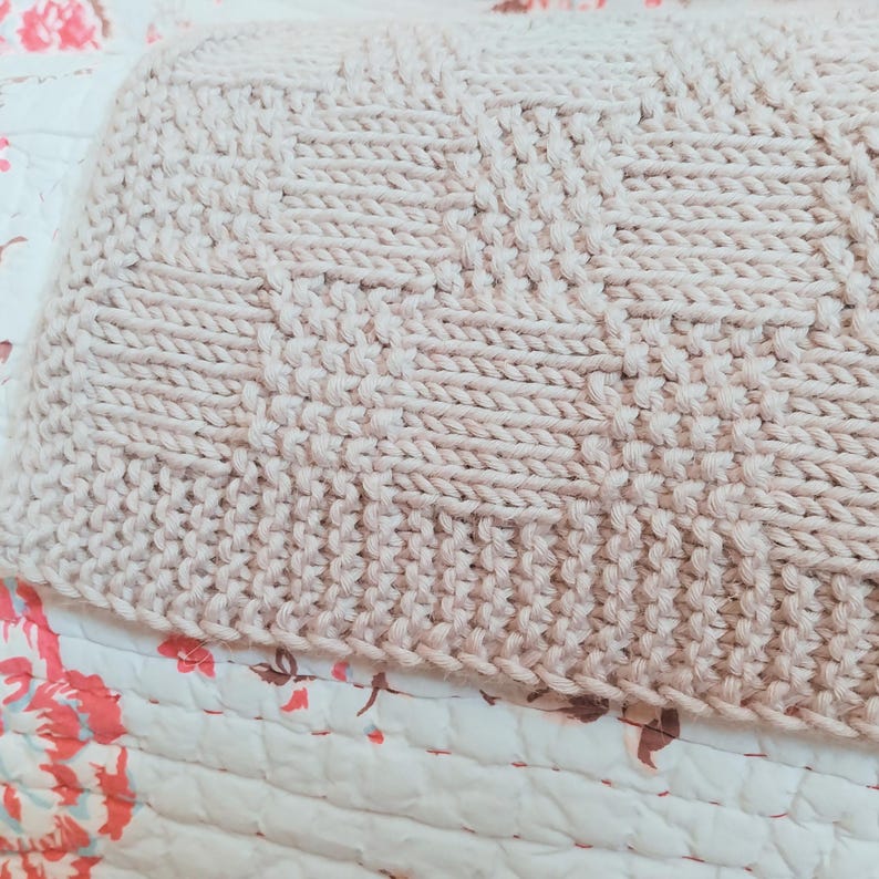May include: Close-up of a beige knitted blanket with a textured pattern. The blanket is made of thick yarn and has a geometric design. The blanket is resting on a floral patterned quilt.