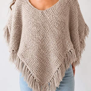 May include: A beige knit poncho featuring a textured, chunky stitch pattern, a wide scoop neckline, and long fringe detailing along the bottom hem. The garment is worn over light blue denim jeans, showcasing a pointed, asymmetrical silhouette.