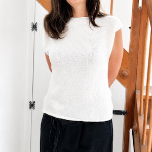May include: A white knit top with a round neckline and short cap sleeves. The garment features a simple, textured stitch pattern throughout, providing a clean and uniform appearance. The top is paired with black trousers featuring a drawstring waist.