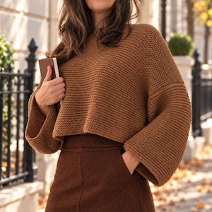 Könnte beinhalten: Ein brauner Strickpullover mit weiter, verkürzter Silhouette und langen, lockeren Ärmeln, kombiniert mit einem dunkelbraunen Rock mit sichtbarer Tasche. Der Pullover hat ein strukturiertes, grobes Strickmuster und einen Rundhalsausschnitt.