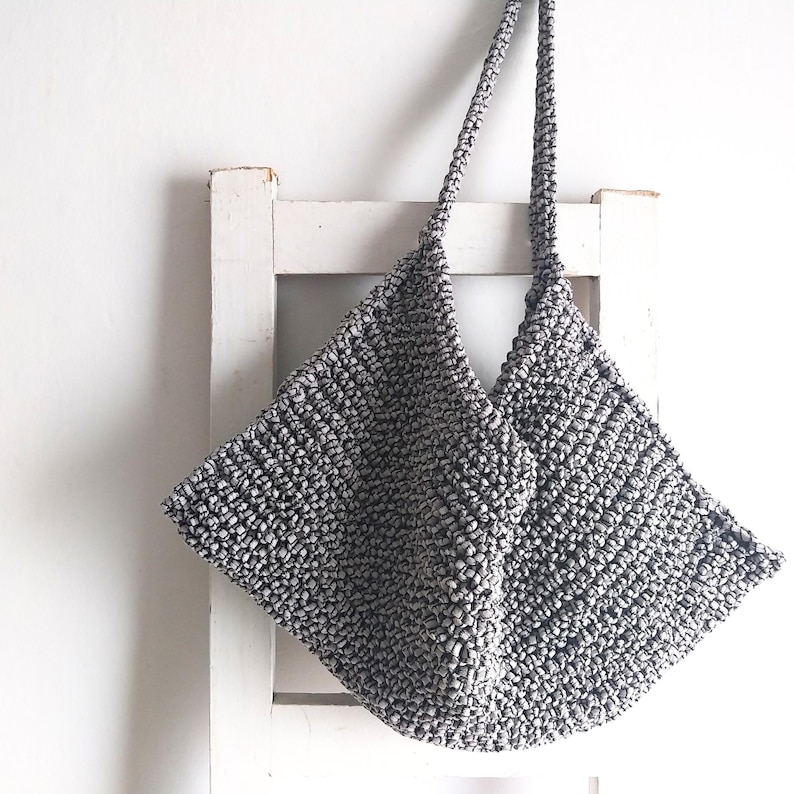 May include: A gray crocheted tote bag with a wide, triangular shape and two long straps. The bag is hanging on a white wooden chair.