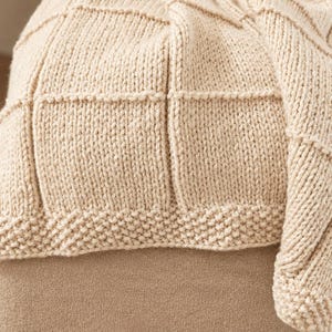 May include: A cream-colored knitted throw blanket featuring a textured grid pattern of raised vertical and horizontal lines. The blanket has a wide border at the bottom edge worked in a garter stitch, creating a distinct contrast to the main body.