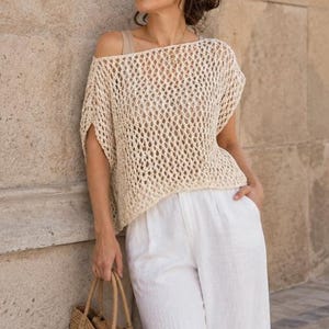 May include: A cream-coloured, loose-fitting crochet top featuring an open-knit diamond pattern, short sleeves, and a wide neckline. The garment is worn over a neutral-toned base layer and paired with white trousers, creating a textured, layered look.