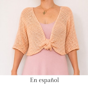 May include: A peach-colored, open-front knit shrug featuring a loose, airy stitch pattern, three-quarter length sleeves, and a front tie closure. The garment is worn over a solid light pink dress, with a gold cross pendant necklace visible.