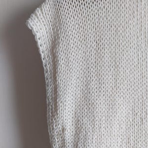 May include: A sleeveless top crafted from off-white yarn in a chunky knit pattern. The garment features a visible stockinette stitch texture with a clean, finished edge along the armhole and a simple, structured neckline construction.