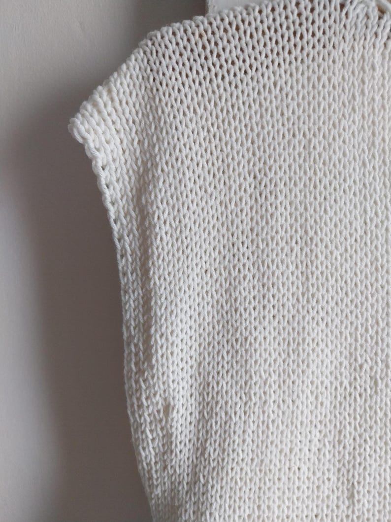 May include: A sleeveless top crafted from off-white yarn in a chunky knit pattern. The garment features a visible stockinette stitch texture with a clean, finished edge along the armhole and a simple, structured neckline construction.