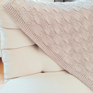 May include: A light beige knitted blanket with a textured, geometric pattern is draped over a stack of white pillows. The blanket's design features a series of rectangular shapes, creating a visually appealing texture. The soft color palette and cozy texture suggest comfort.