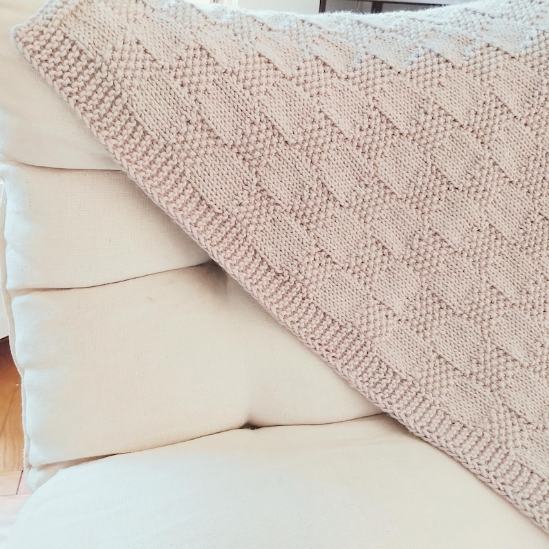 May include: A light beige knitted blanket with a textured, geometric pattern is draped over a stack of white pillows. The blanket's design features a series of rectangular shapes, creating a visually appealing texture. The soft color palette and cozy texture suggest comfort.