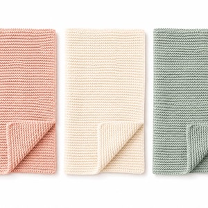 May include: Three rectangular knitted blankets arranged side by side. The blankets feature a consistent garter stitch texture and are colored in soft shades of dusty pink, cream, and sage green, each with one corner folded over to show the reverse.
