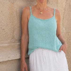 May include: A light turquoise sleeveless knit tank top with thin shoulder straps and a scoop neckline, paired with a white skirt featuring a drawstring waist. The tank top has a simple, smooth texture and a relaxed fit, providing a clean look.