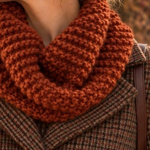 Peut inclure: Une écharpe infinie en tricot épais de couleur orange brûlé avec une texture en point mousse prononcée. L'écharpe est enroulée en double boucle autour du cou et portée sur un manteau en laine à motif pied-de-poule marron et noir.