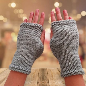 Op de afbeelding: Een paar grijze vingerloze handschoenen van gebreid garen. De handschoenen worden getoond op een houten oppervlak.