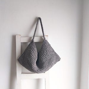 May include: A gray crocheted tote bag with a wide, open top and two long handles. The bag is hanging on a white wooden ladder.